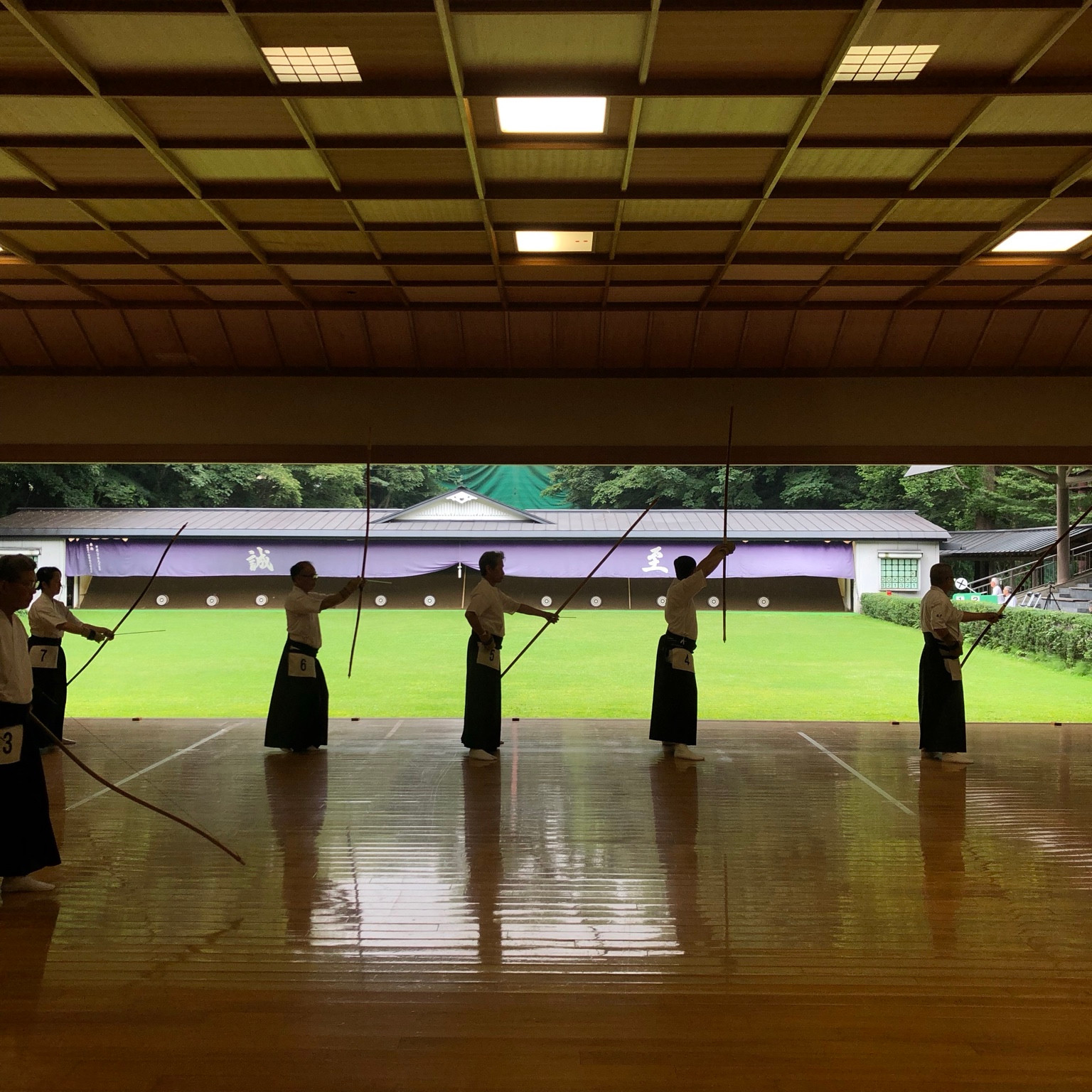 Archers stand in a row holding bows in a covered kyudo hall, facing the outdoor range.
