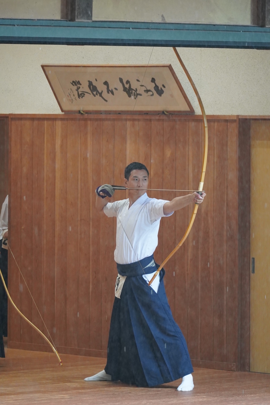 Kensuke Ueoka holding a drawn Japanese bow inside a wooden kyudo hall.